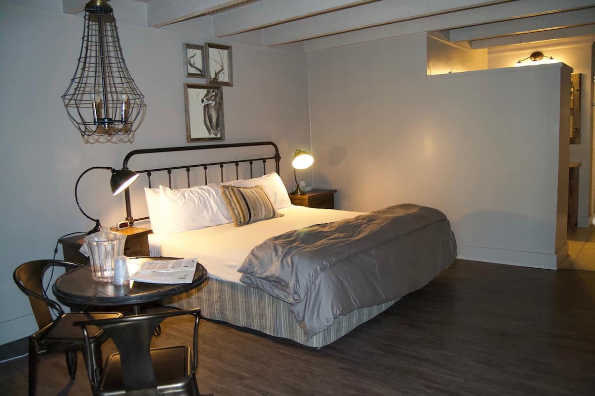 A neatly arranged bedroom with a bed, bedside tables, a lamp, a chair, and a small table with a glass container and papers.