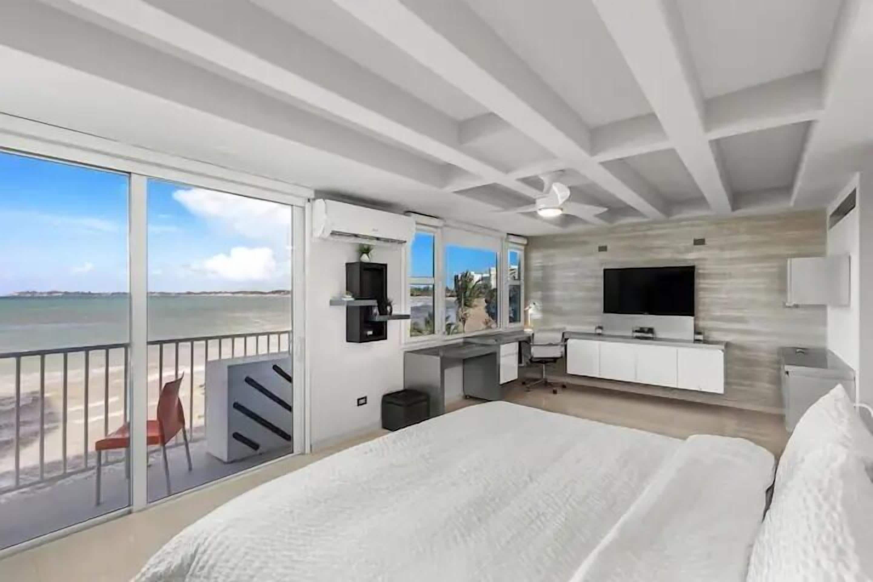 A modern hotel room with a large bed, a desk with a TV, and a balcony with a view of the beach.