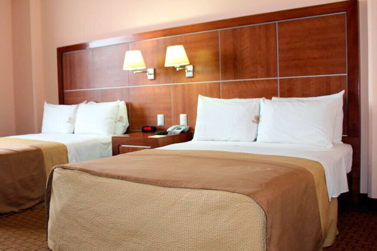 Habitación de hotel con dos camas, cabecero de madera, lámparas de noche y mesitas de noche con un reloj y un teléfono.