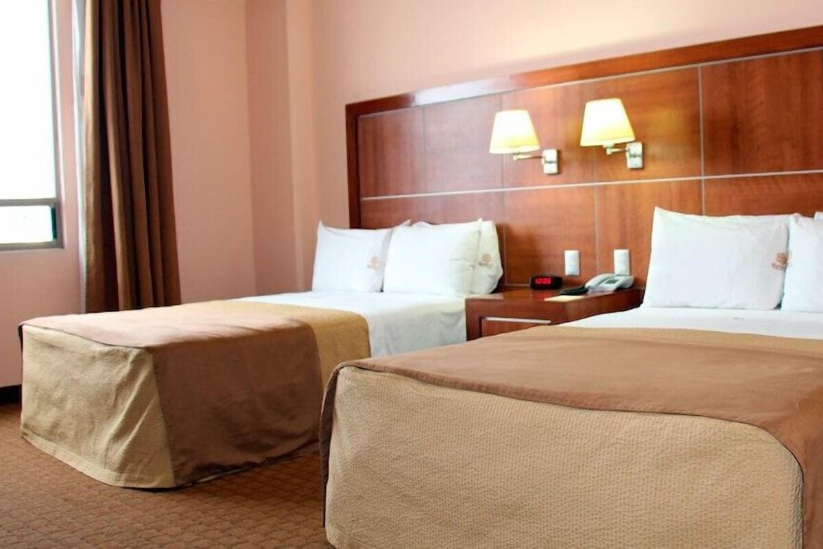 Une chambre d’hôtel avec deux lits, une tête de lit en bois, des lampes de chevet et une fenêtre avec des rideaux.