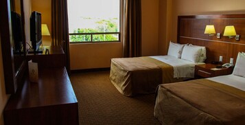 Habitación de hotel con dos camas, un escritorio con televisor, una ventana con cortinas y dos lámparas fijadas a la pared.