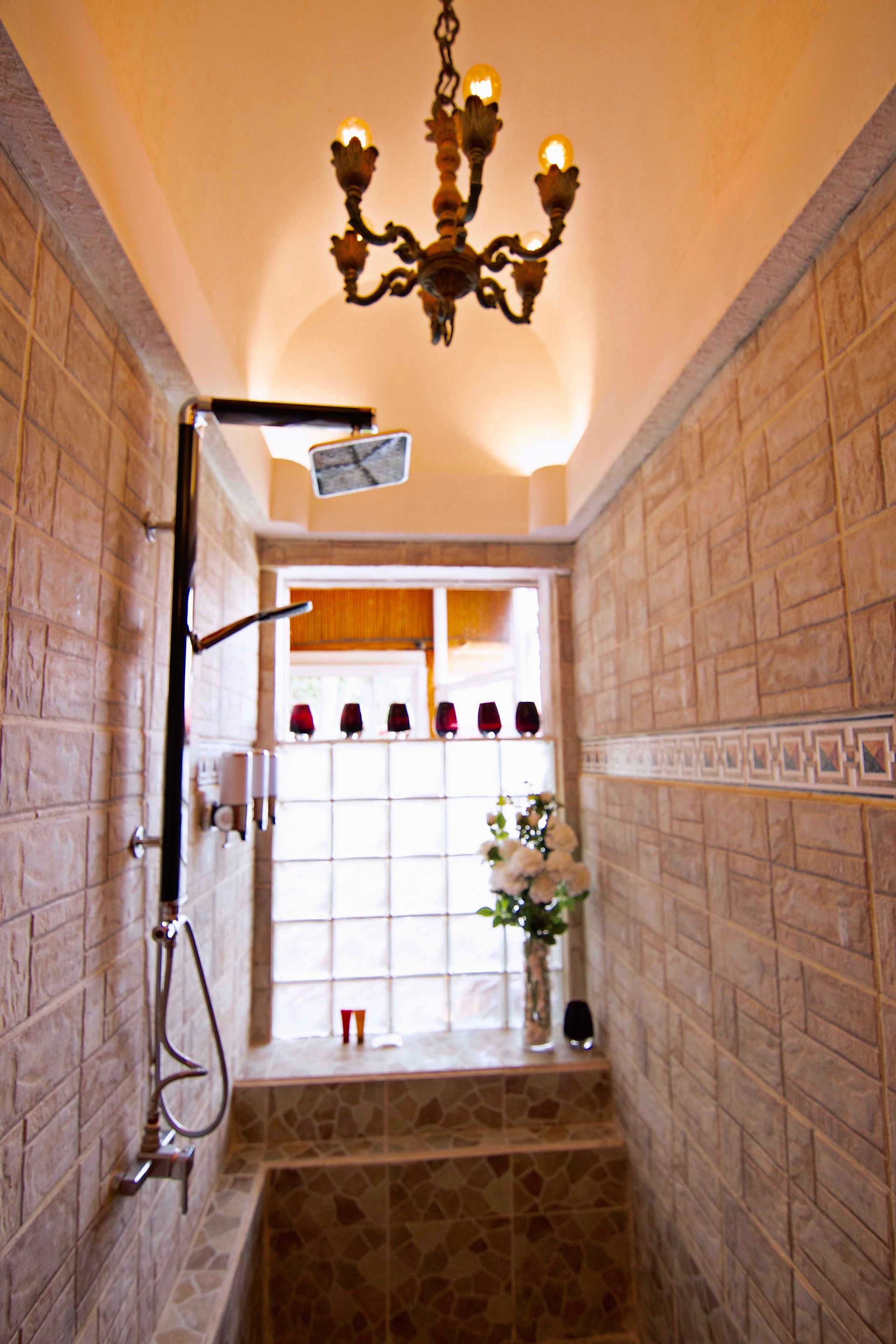 A bathroom with a shower, a wall-mounted mirror, a chandelier, and a window with decorative items.