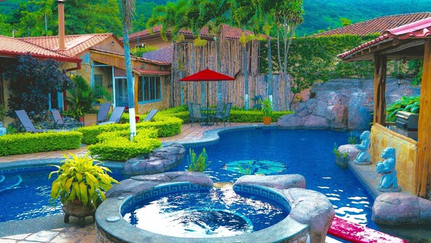 A swimming pool with a stone fountain, surrounded by tropical plants and a wooden fence. - Angel's Cave (Escazu)