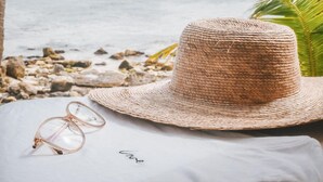Un sombrero de paja y gafas sobre una superficie blanca con la palabra 'love' escrita, con un fondo de playa que incluye palmeras y el océano.