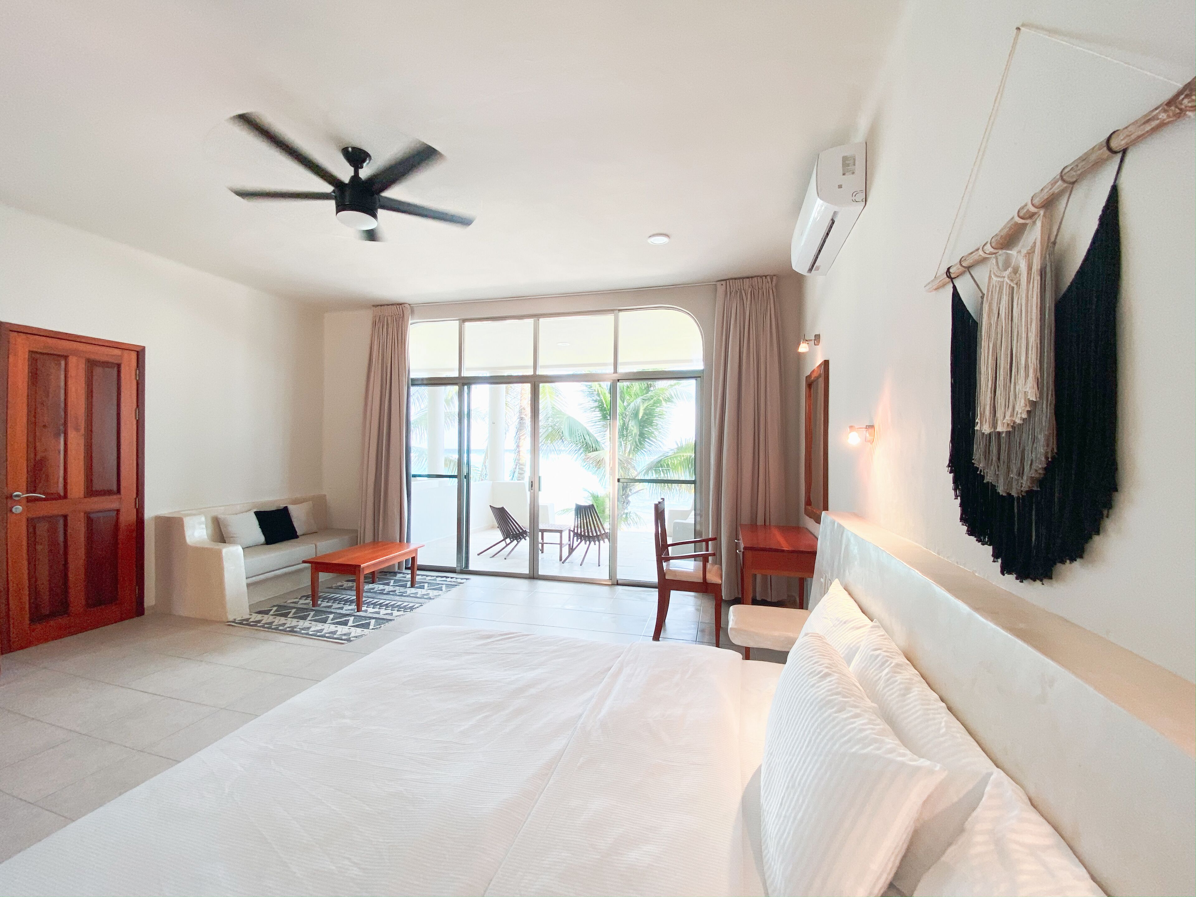 Une chambre d’hôtel avec un grand lit, un ventilateur de plafond, une porte-fenêtre coulissante donnant sur un balcon équipé de chaises, et une porte en bois.