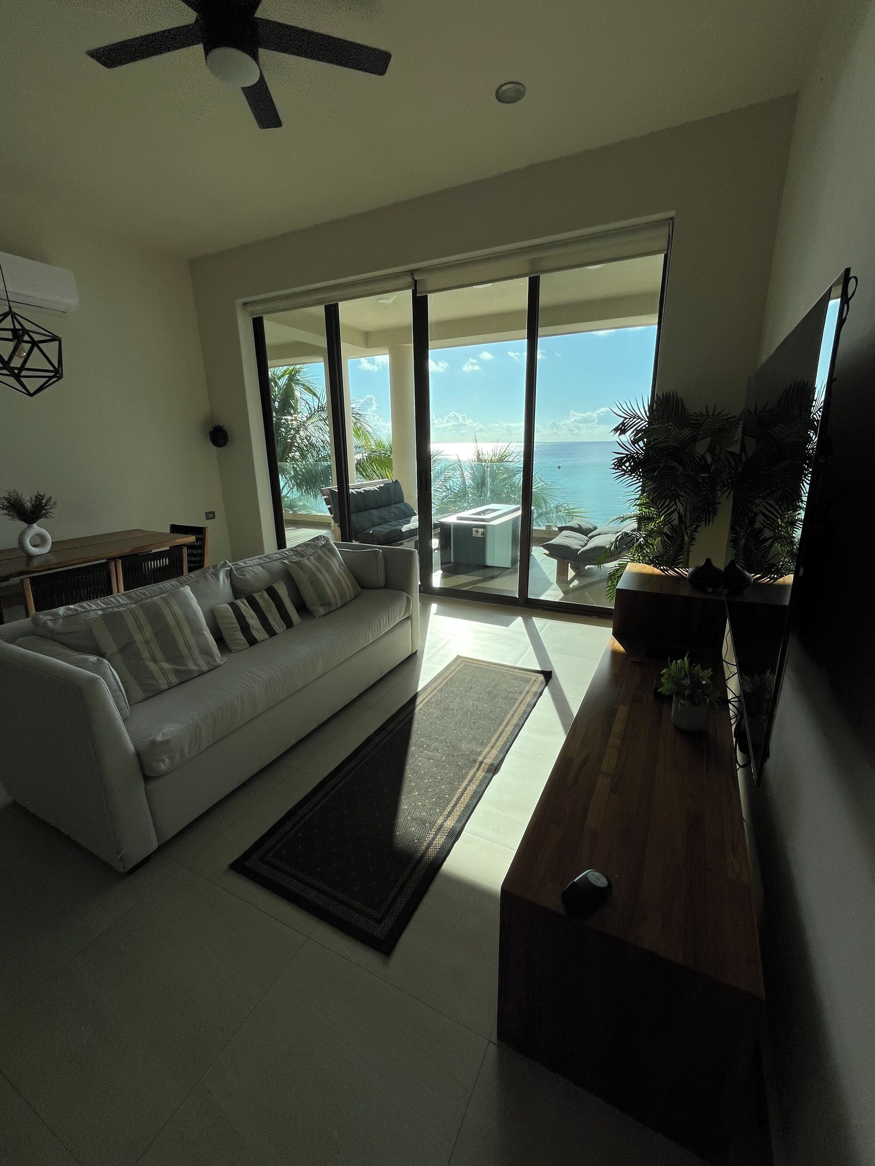 A modern living room with a sofa, a coffee table, and a TV. There is a large sliding glass door leading to a balcony with a view of the ocean.