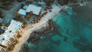Vista aérea de un resort en la playa con cabañas de techo de paja, palmeras y una piscina.