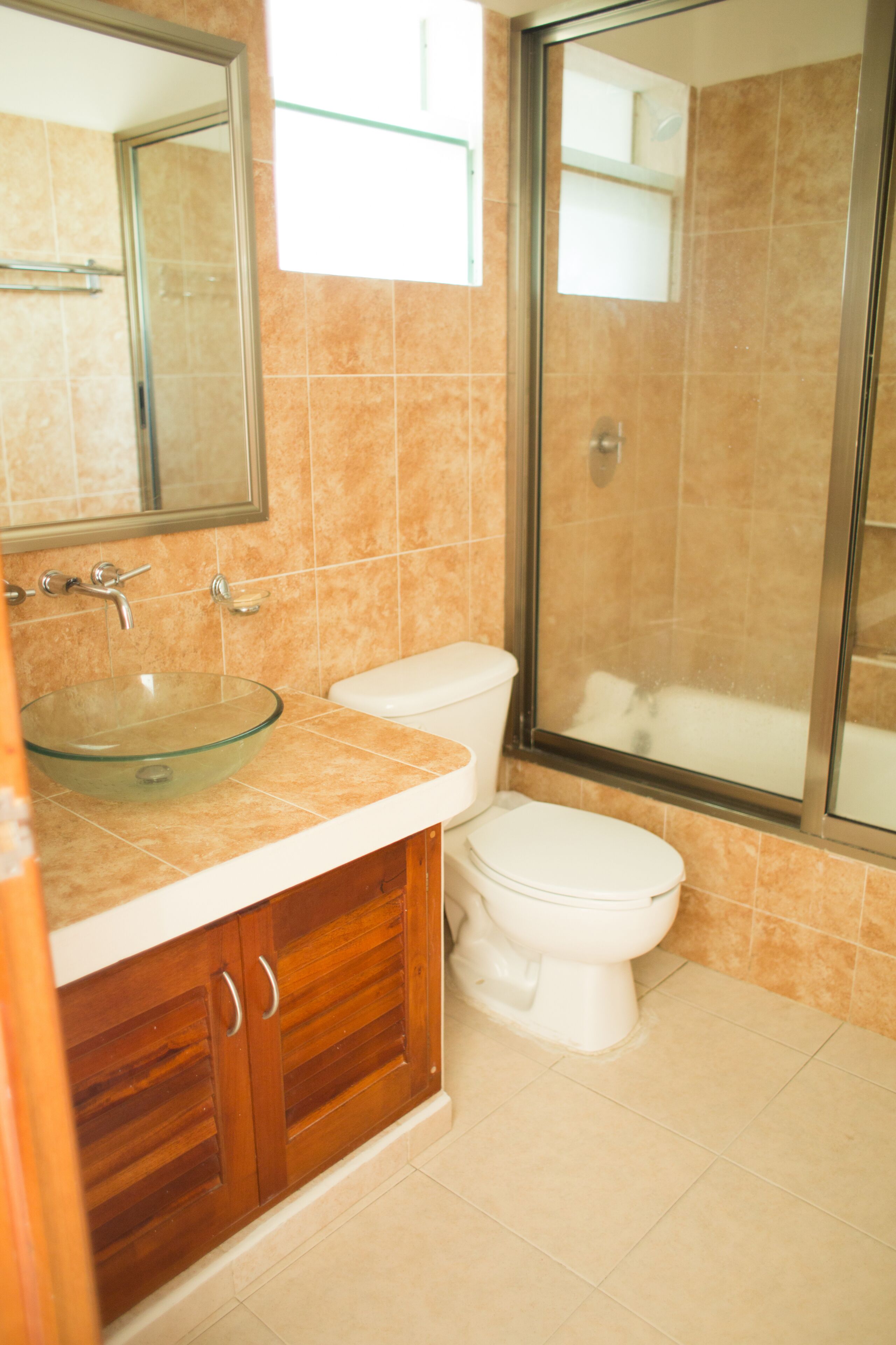 Un baño con lavamanos de vidrio, muebles de madera, inodoro blanco y cabina de ducha con paredes de vidrio.