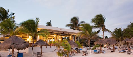 A beachside resort with thatched umbrellas, sun loungers, and a building with large windows.
