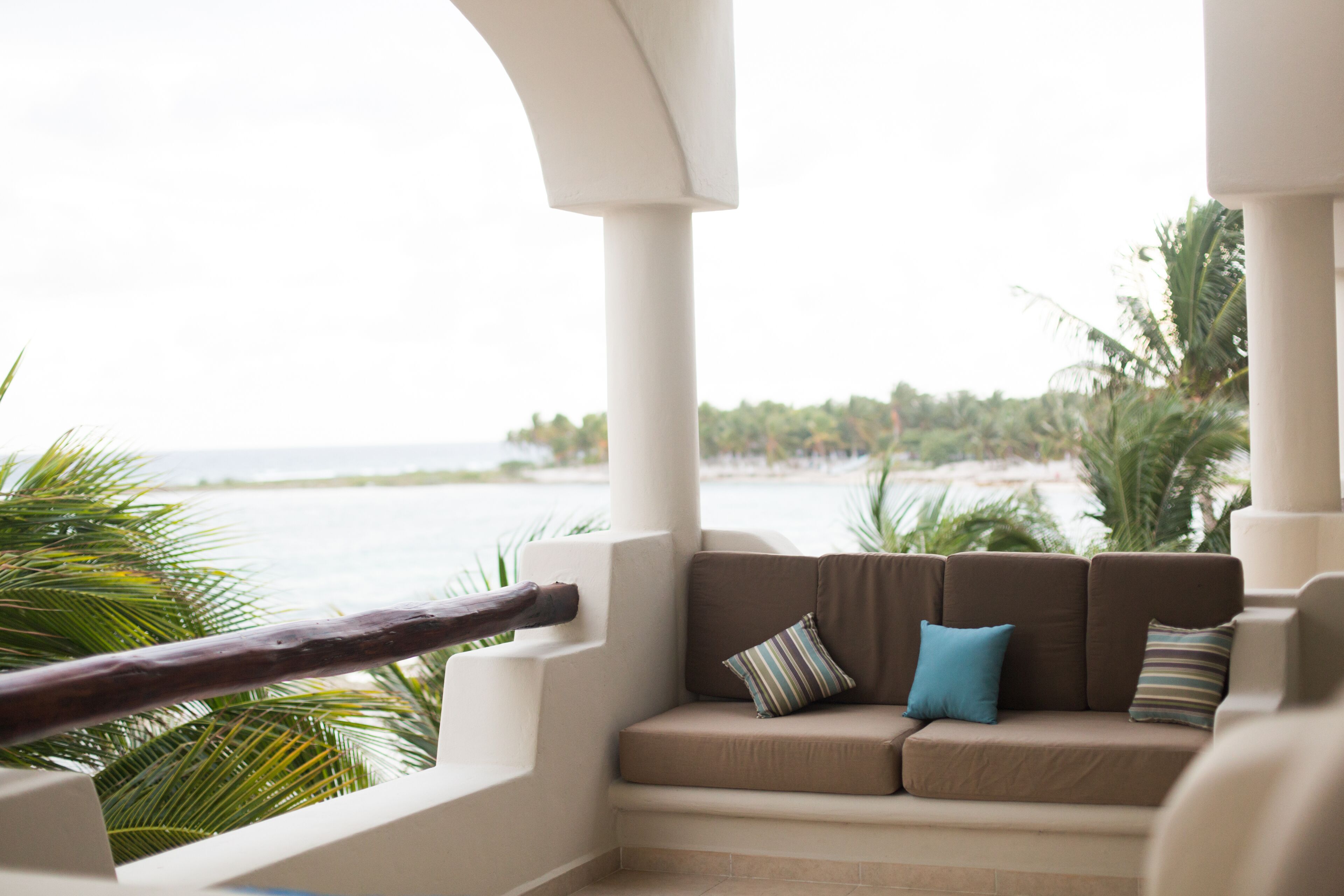A balcony with a cushioned seating area, a view of the ocean, and palm trees.