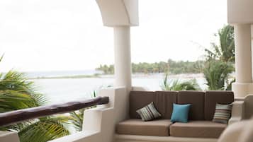 A balcony with a cushioned seating area, a view of the ocean, and palm trees.