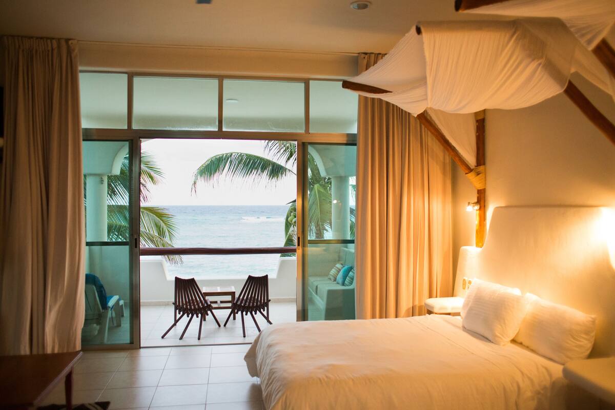 A bedroom with a large bed, a balcony with a view of the ocean, and a table with chairs.