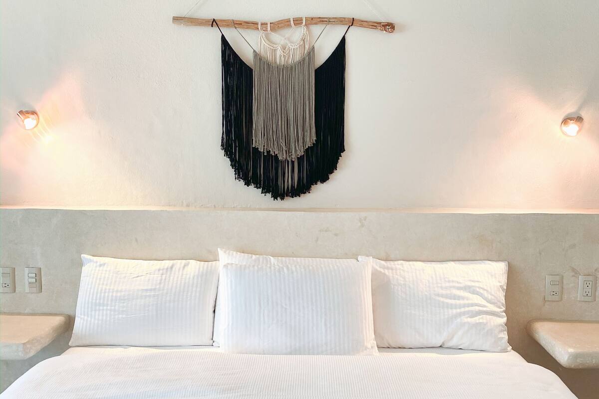 A bed with white linens, a macrame wall hanging, and two bedside lamps.