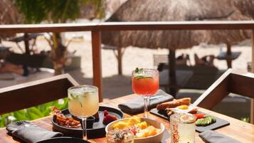 Uma mesa de madeira com diversos pratos e bebidas, com o cenário de uma praia, toldos de palha e palmeiras.