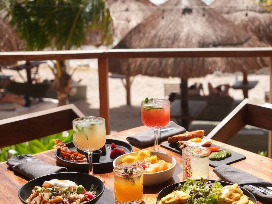Uma mesa de madeira com diversos pratos e bebidas, com o cenário de uma praia, toldos de palha e palmeiras.