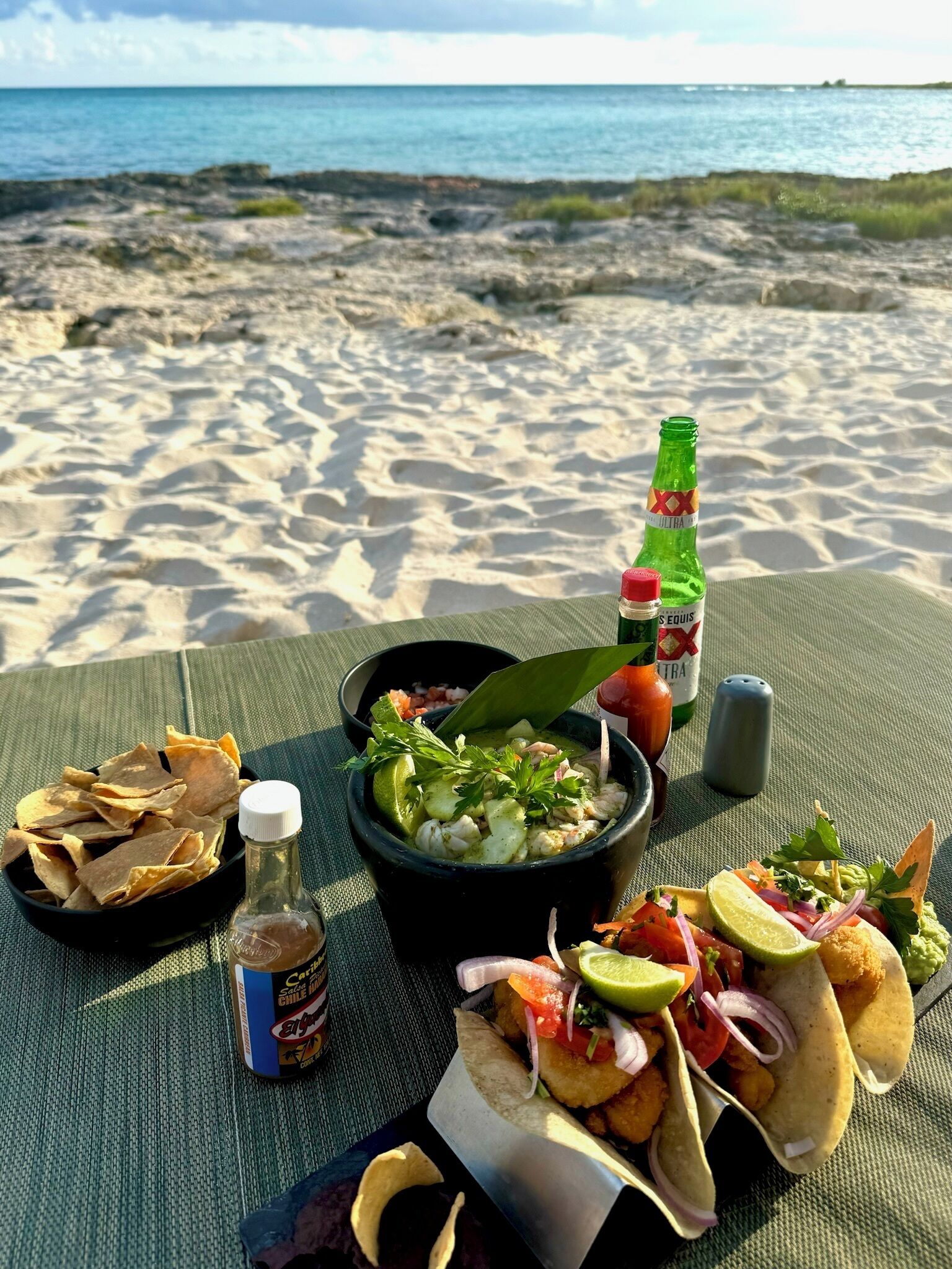 A beachside meal setup with a bowl of soup, chips, a bottle of sauce, and a drink, with the ocean in the background.