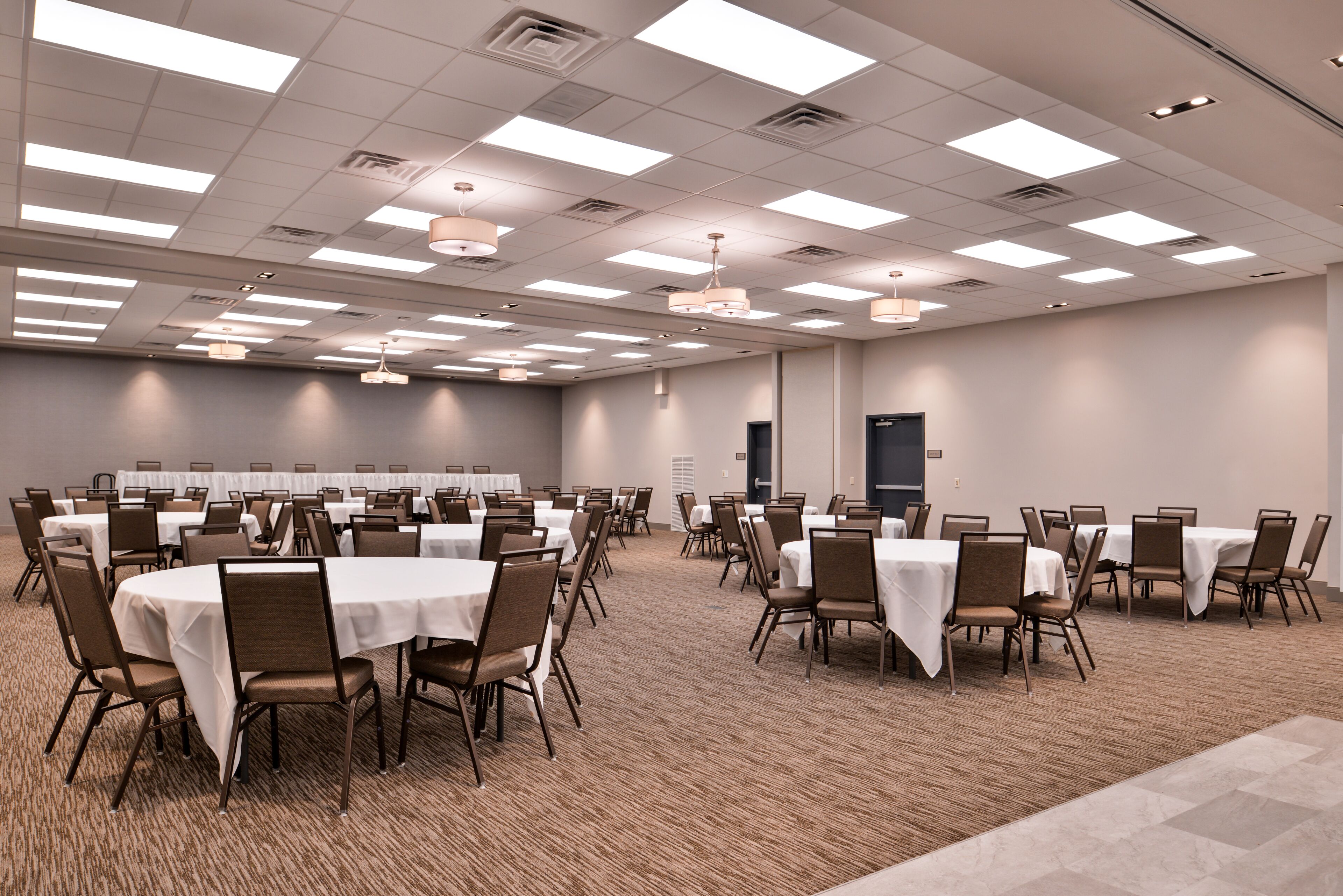 A large conference room with multiple tables and chairs, arranged in rows, and equipped with ceiling lights and ventilation.