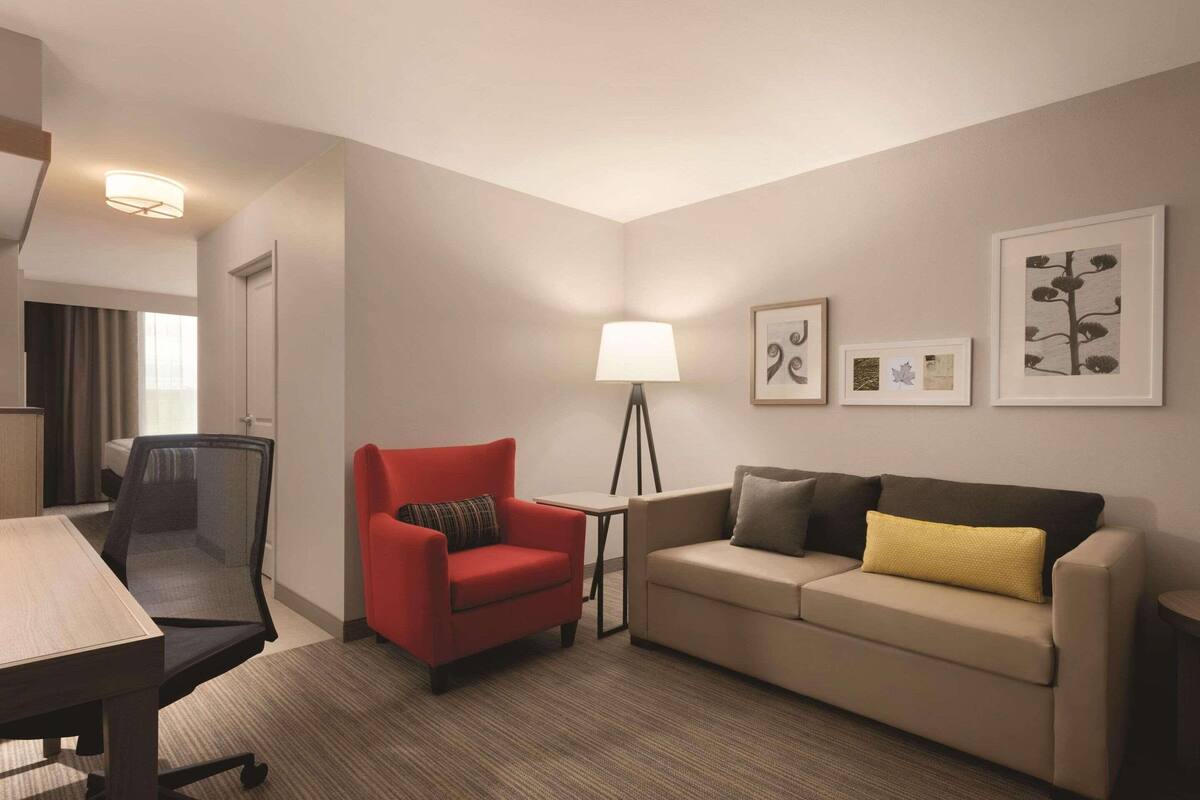 A hotel room with a desk, a red armchair, a beige sofa, a floor lamp, and framed artwork on the wall.