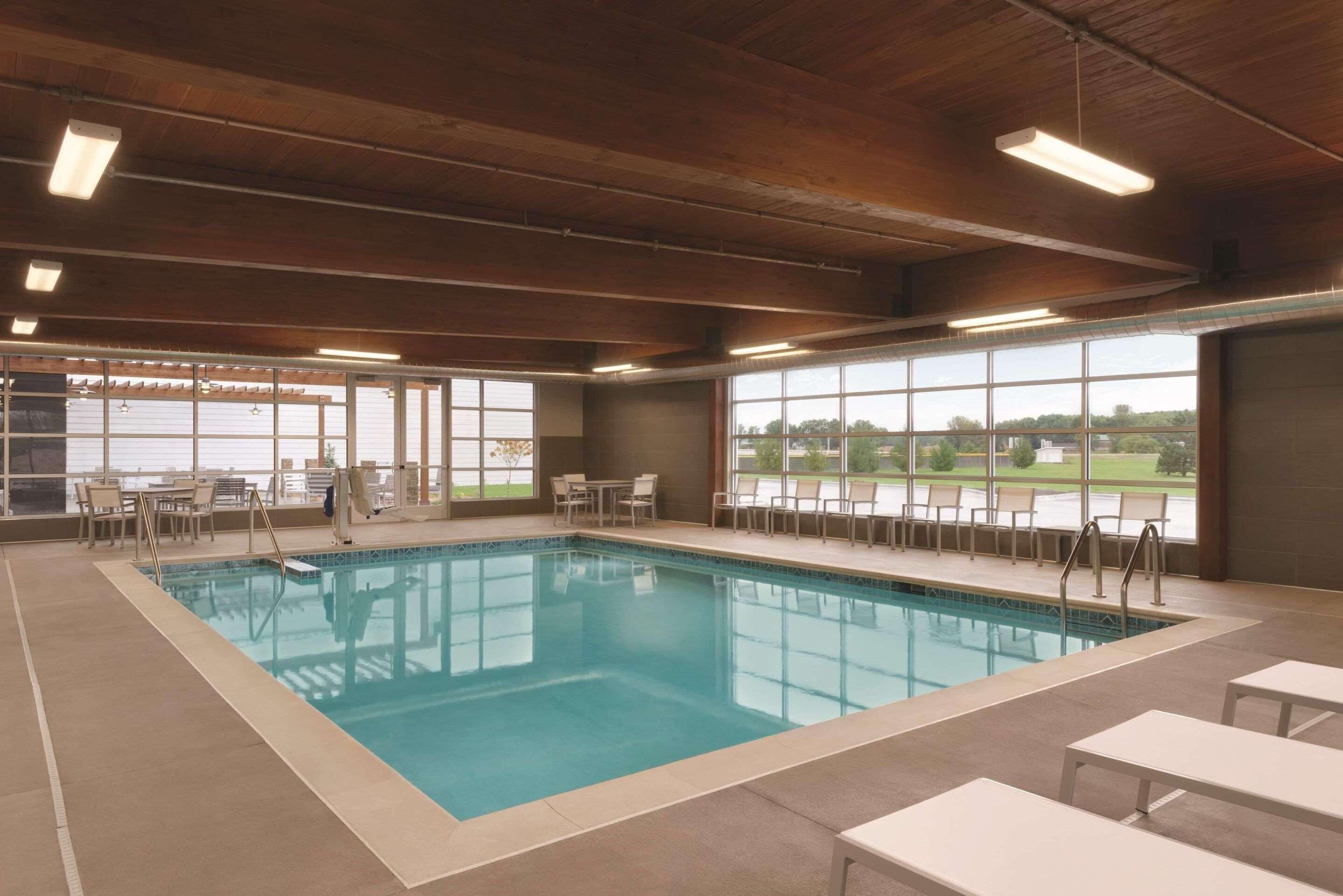 An indoor swimming pool with clear blue water, surrounded by lounge chairs and tables, with large windows offering views of the outdoors.