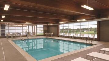 An indoor swimming pool with clear blue water, surrounded by lounge chairs and tables, with large windows offering views of the outdoors.
