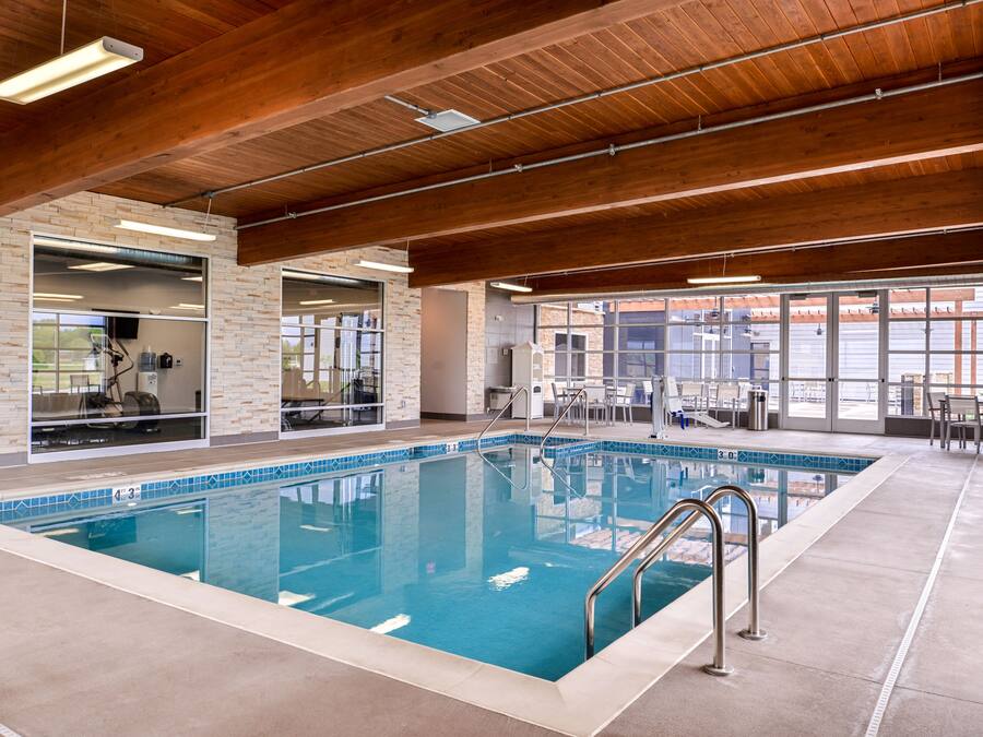 An indoor swimming pool with a clear view of the surrounding area, including a brick wall, a glass door leading to a room, and a seating area with tables and chairs.