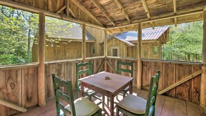 Un porche cubierto de madera con una mesa y sillas, ventanas grandes y vistas a una casa y árboles.