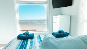 A bedroom with a large window overlooking a beach, a white dresser, and two folded towels on the bed. - Summer Wind, Beachfront with incredible views! 5 miles from Galveston sea wall! (Galveston)