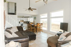 A spacious living room with a sectional sofa, a dining area with wooden chairs, a kitchen in the background, and large windows offering a view.