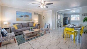 A living room with a gray sofa, patterned armchairs, a coffee table, and a dining area with yellow chairs and a table set for four. - Beautiful Apartment @ The Botanical Gardens #6 (San Antonio)