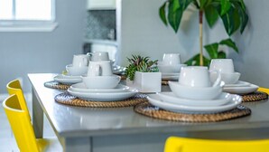 A neatly set dining table with white plates, bowls, and cups, accompanied by woven placemats and a small potted plant. - Beautiful Apartment @ The Botanical Gardens #6 (San Antonio)