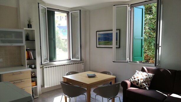 A small, well-lit room with a dining table and chairs, a sofa, and two windows with open shutters. - Pisolo home holidays near city center and Sant'Orsola Hospital (Bologna)