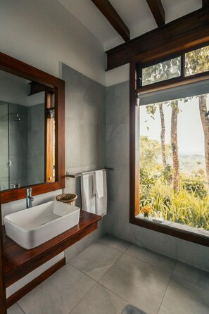 A bathroom with a large mirror, a white sink, and a window with a view of trees and greenery.