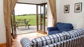 Un salon avec un canapé à rayures, un fauteuil bleu et une porte en verre donnant sur un espace extérieur équipé d’une table de pique-nique.