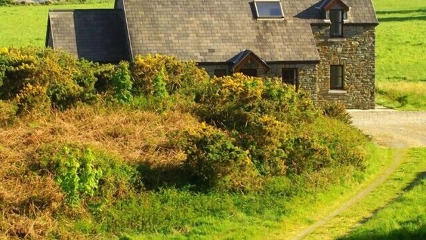 Une maison en pierre, dotée d’un toit en pente et d’une petite dépendance attenante, entourée de champs verdoyants et offrant une vue dégagée sur la mer.