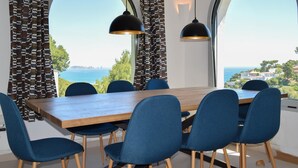 A dining area with a wooden table and blue chairs, large windows with patterned curtains, and pendant lights.