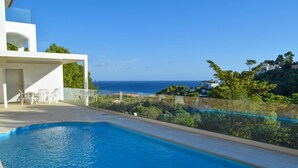 A swimming pool with a view of the sea, surrounded by greenery and a white building.