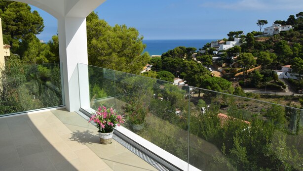 A balcony with a potted plant, overlooking a coastal town with houses and greenery.