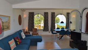 A living room with a blue sofa, a dining table with chairs, a TV, and a view of the sea through large windows.