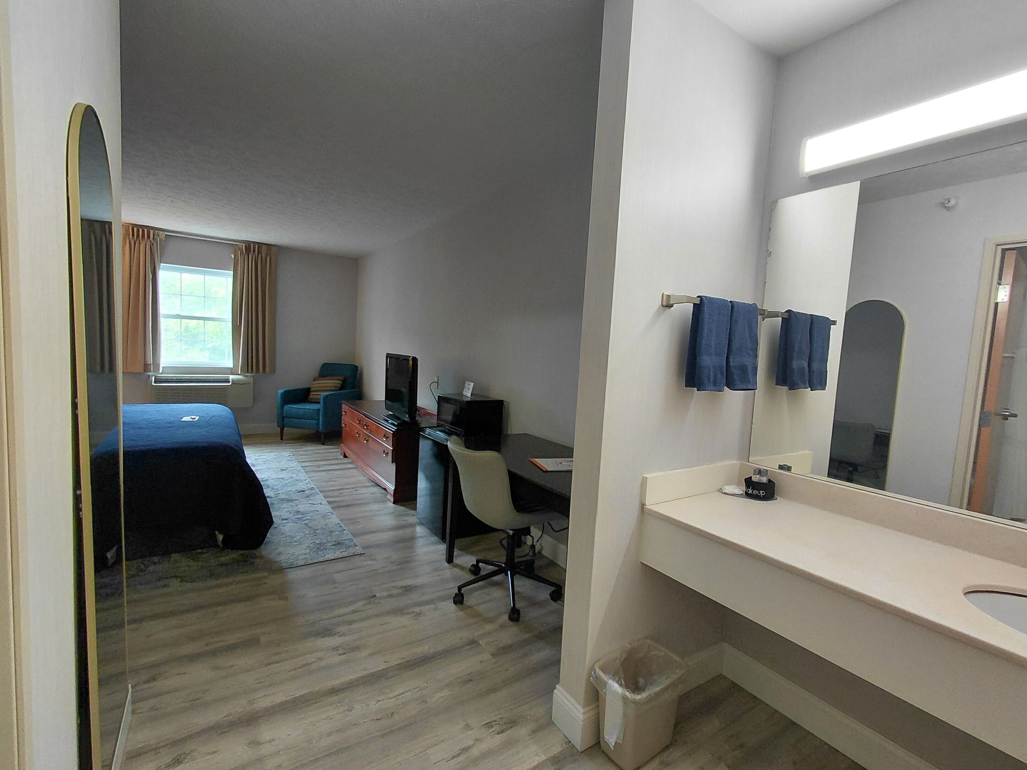 A hotel room with a bed, a desk with a computer, a chair, a small trash bin, and a bathroom with a sink and mirror.