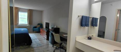 Habitación de hotel con cama, escritorio con computadora, silla, basurero pequeño y baño con lavamanos y espejo.