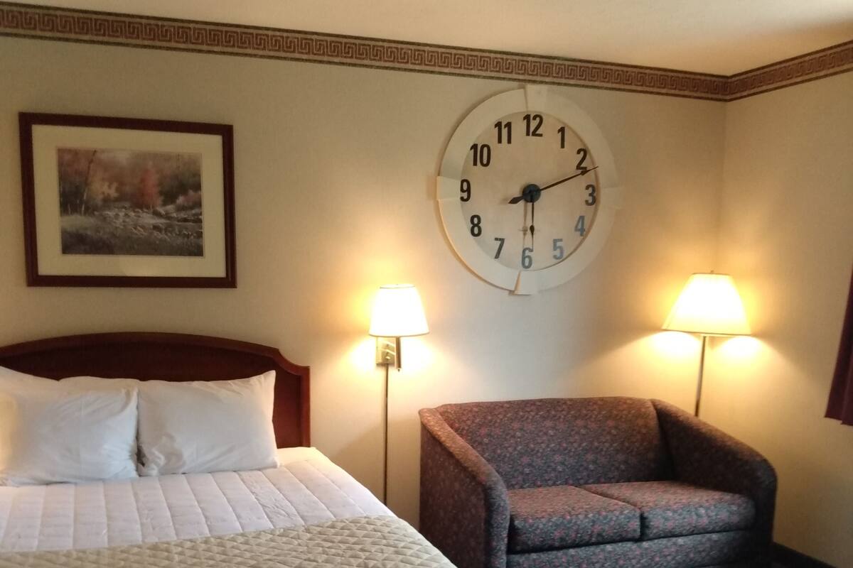 A hotel room with a bed, a chair, two wall lamps, and a clock.