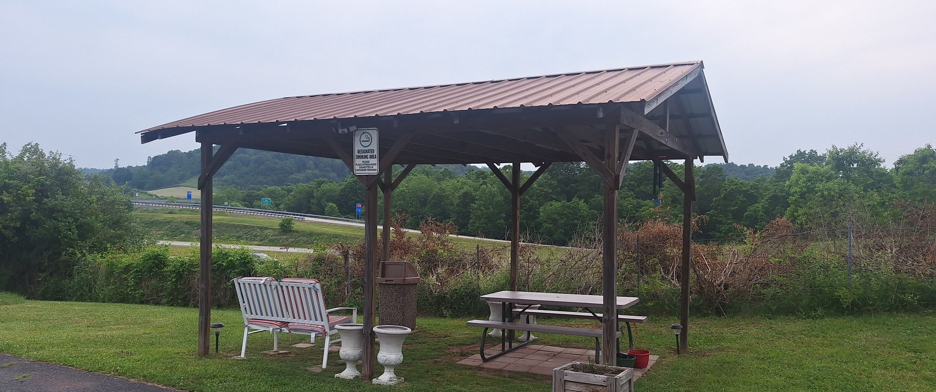 A gazebo with a picnic table and benches, surrounded by greenery and a road in the background.
