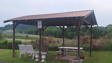 A gazebo with a picnic table and benches, surrounded by greenery and a road in the background.