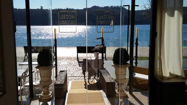 A view from a hotel room with a glass door, overlooking a beach and a lake. There are two signs reading "HOTEL" and "HOTEL" on the glass door. - Duomo Hotel (Toscolano Maderno)