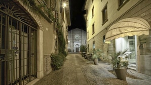 A narrow cobblestone street with buildings on either side, a gate on the left, and a church with a bell tower in the background. - Duomo Hotel (Toscolano Maderno)