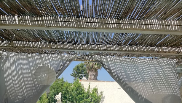 A pool area with a clear blue pool, surrounded by a pergola with a view of a palm tree and a statue. There is a round table with chairs in the foreground. - Villa 'Oasis' with exclusive pool (Siracusa)