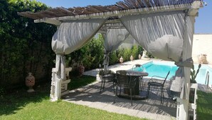 A covered outdoor seating area with a pool, surrounded by greenery and decorative pots. - Villa 'Oasis' with exclusive pool (Siracusa)