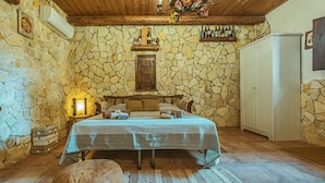 A rustic bedroom with a stone wall, wooden ceiling, a bed with a striped bedspread, a wooden bench, a small table with a lamp, and a wardrobe. - Villa 'Oasis' with exclusive pool (Siracusa)