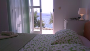 A bedroom with a bed, a window with a view of the sea, a bedside table with a lamp, and a towel on the floor. - Romantic, quite, seafront apartment, sunbeds, barbecue, all by the sea. For 2p (Komarna)