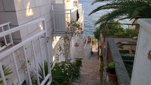 A narrow outdoor corridor with a stone floor, bordered by white railings and potted plants. - Romantic, quite, seafront apartment, sunbeds, barbecue, all by the sea. For 2p (Komarna)